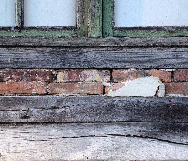 An upclose photo of an aged and weathered wall. Thin, discoloured briks are in the centre on the picture, with ancient-looking wooden beams below and above. The top quarter of the photo shows a worn green window frame and milky white coloured windows.
