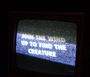 A photo of a tv up-close in a darkened room. There is static on the screen and the words "join the wind up to find the creature" displayed in block white capitals on the screen.