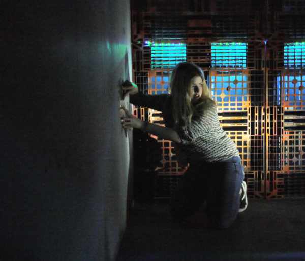 A photo of a person crouching down next to a wall while holding something against it. They are looking backward over their shoulder. A grey wall fills the left hand side of the picture where the person is crouched. The rest of the picture shows an orange industrial-looking wall backlit with light blue lighting.