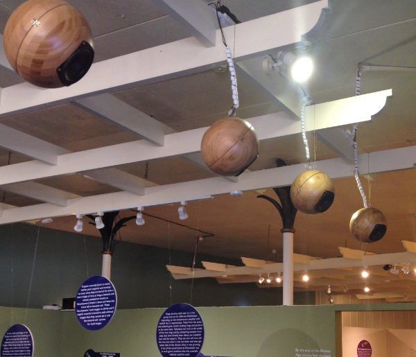 Four wooden spheres have been adapted to hold speaker cones. They are hanging from the ceiling. Unreadable museum information signs also hang.