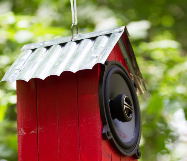 A picture of a red birdbox with a corrigated gable tin roof hanging in the woods. A speaker has been put in the front of the birdbox. 10 of these were hung around a section of woodland to create a magical surround-sound effect.